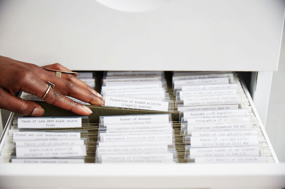 A well-manicured hand with brown skin rifles through folders in a filing cabinet. Their fingers linger on a file labeled, "Proportion of traded wildlife illegally poached."
