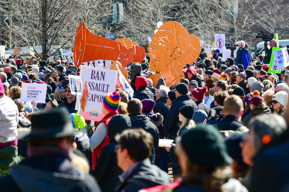 In an outdoor crowd dressed in coats and hats stands a large orange cut-out of a person shouting into a megaphone. News cameras also appear in the crowd, and someone's sign reads BAN ASSAULT WEAPONS.