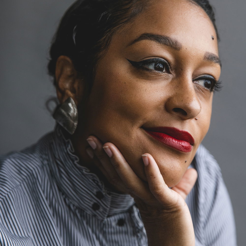 Eve, a light skinned Black woman, leans forward, smiling slightly and looking off camera. Her dark brown hair is braided close to her head and she has on bright red lipstick and a high-collared button-up shirt. She is wearing winged eyeliner and has freckles.