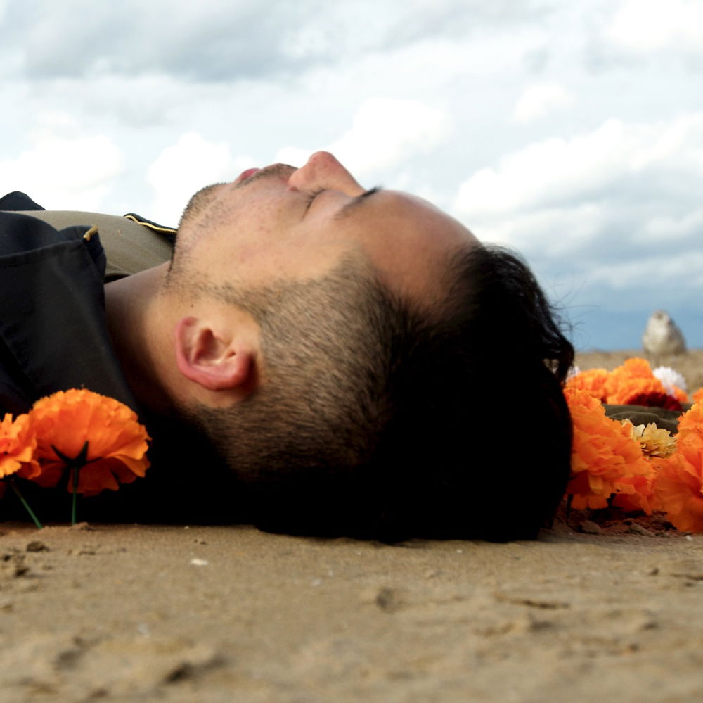 A young Asian American man dressed in work clothes but without shoes, lies facing up on the beach, hands folded on his abdomen, surrounded by orange and yellow marigolds.