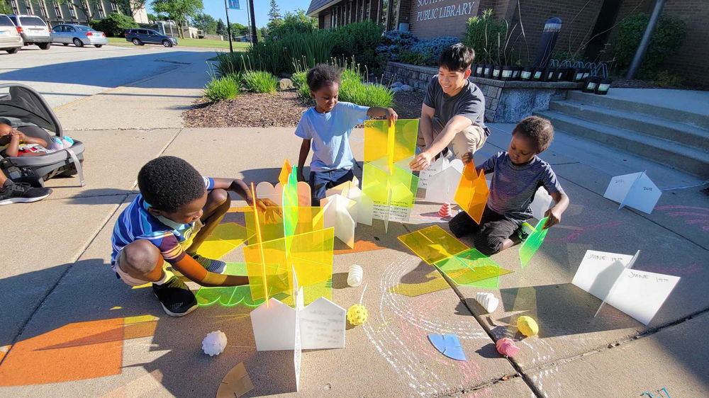 A group of four kids are crouched down on the pavement as they play with neon yellow and green acrylic sheets to create custom shapes.