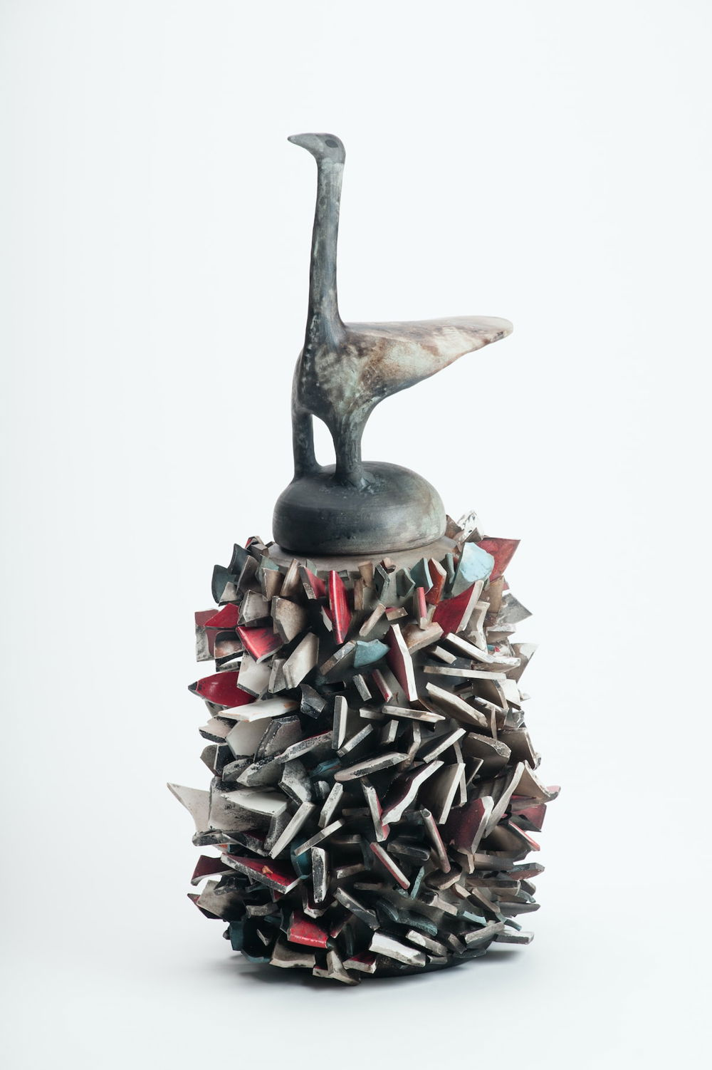 An abstract grey stone sculpture resembling a bird with a beaked head, long neck, oblong triangular body, and two-cylinder shaped legs. The sculpture stands on a circular pedestal that is covered in jagged ceramic shards.