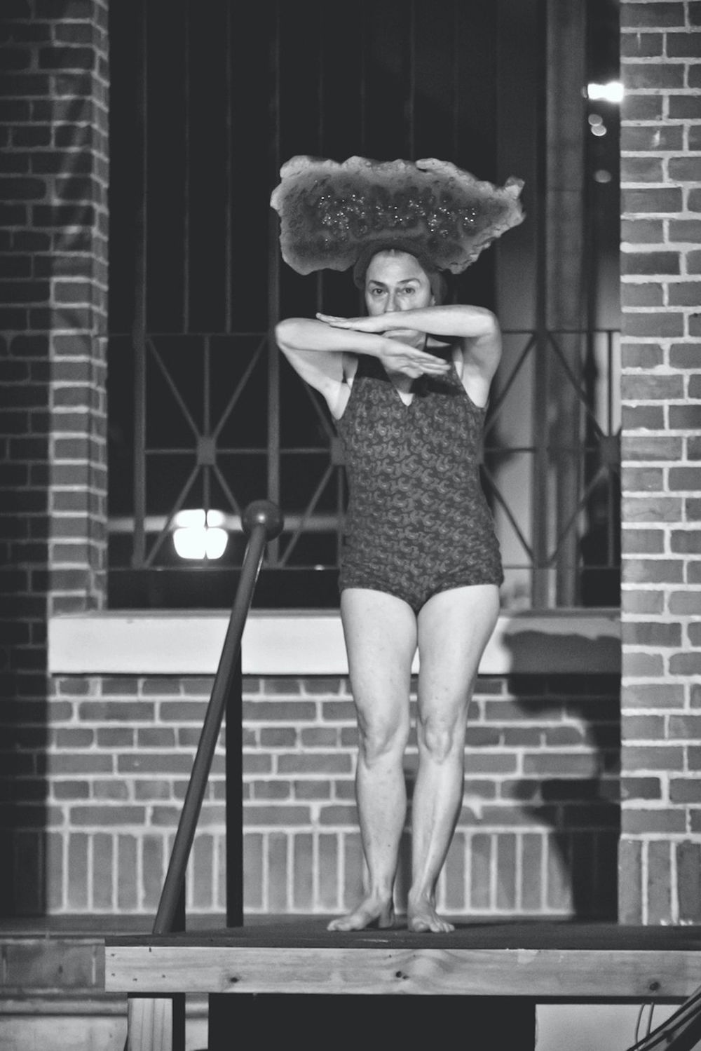 A black-and-white photograph of a woman in a leotard standing on a wooden ledge in front of a brick building; her arms are folded in front of her mouth and she looks out over them. She wears a headpiece that is shaped like the island of Puerto Rico.