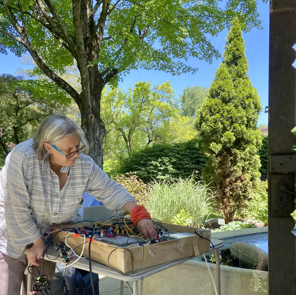 Liz Phillips adjusts patch cables on a medium-sized synthesizer in a brightly sunlit outdoor area surrounded by trees and foregrounded by an old wooden fence.