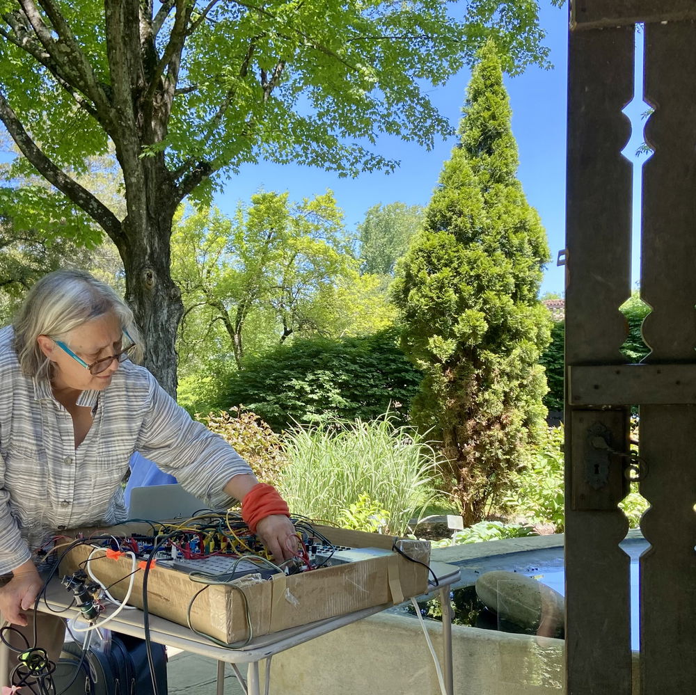 Liz Phillips adjusts patch cables on a medium-sized synthesizer in a brightly sunlit outdoor area surrounded by trees and foregrounded by an old wooden fence.