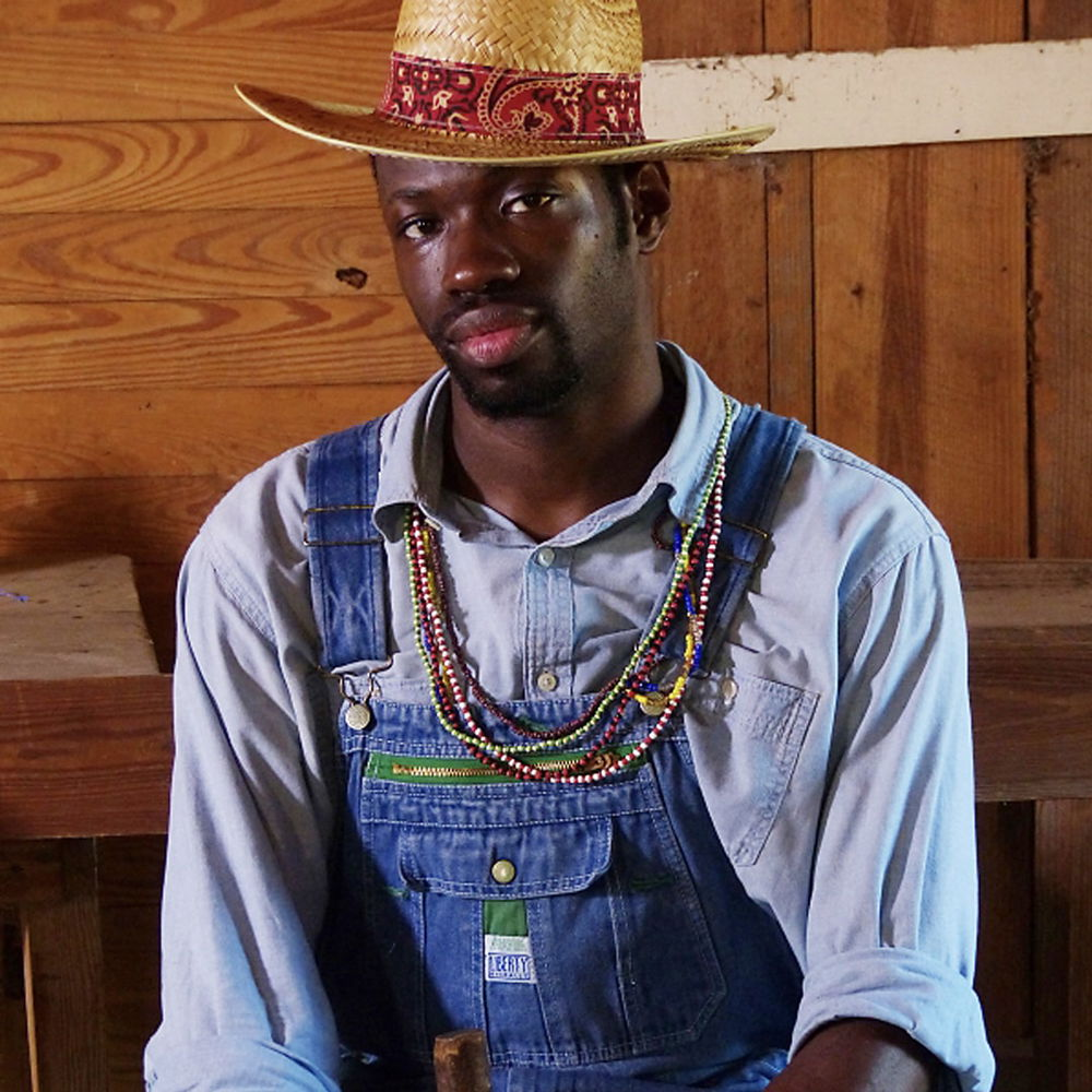 A black man sits against a wooden wall, gazing into the camera. He is wearing a straw hat at an angle, a collared shirt under denim overalls, and colorful beaded necklaces.