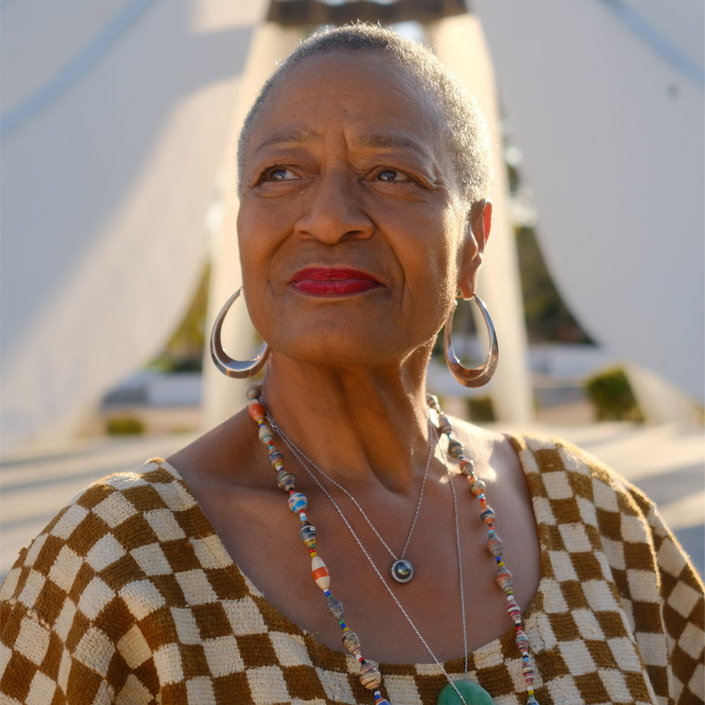 A Black woman in her early seventies looks out beyond the camera. She wears silver hoop earrings, red lipstick, and a checkered top with a paper beaded necklace and a jade pendant. The white, angular planes of the King Memorial sculpture in Compton, CA, rise up behind her.