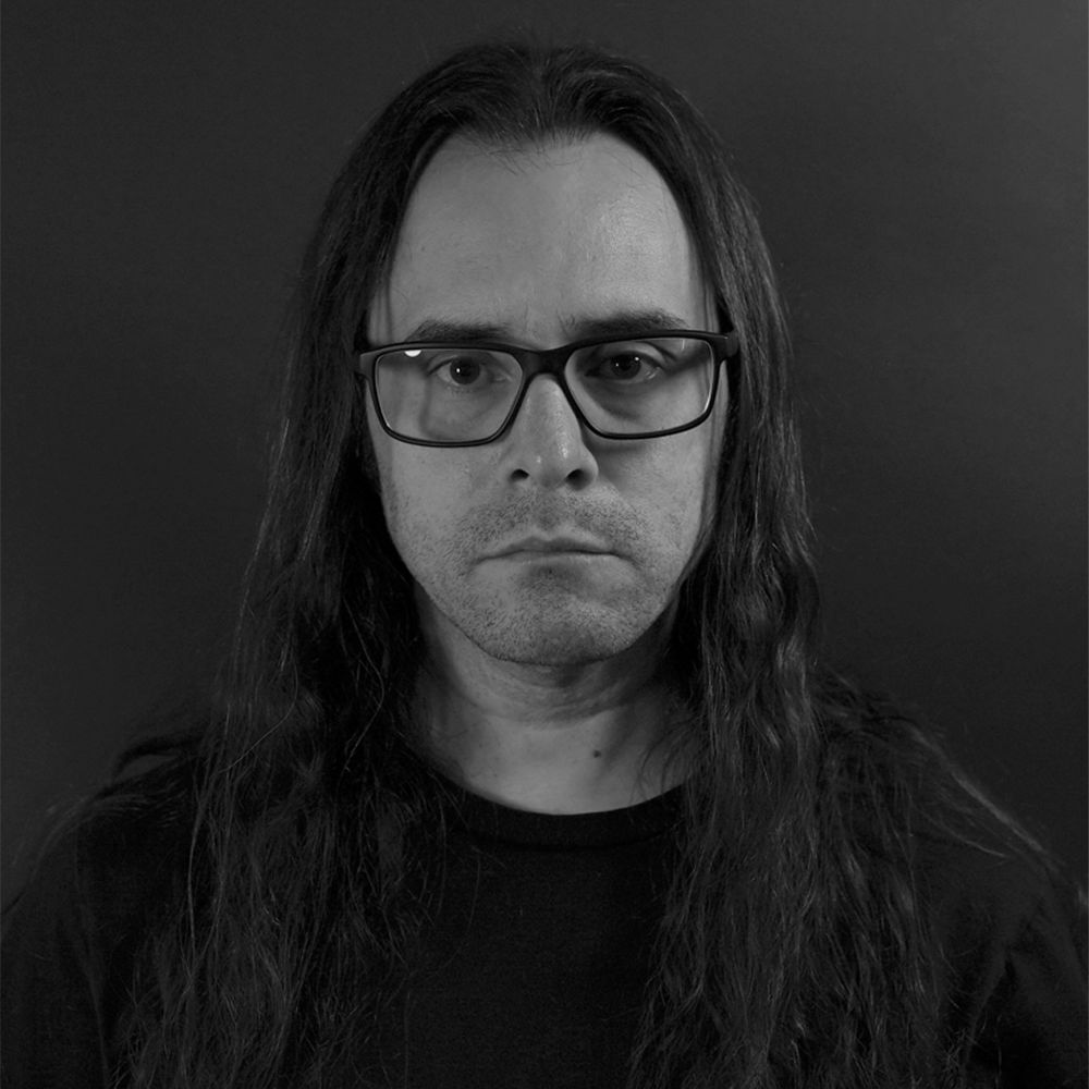A grayscale photograph of a man with long dark hair wearing glasses. The photograph has little light, which causes his dark hair and black shirt to fade into the background.