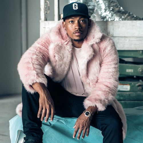 A portrait of a man sitting in an empty showroom, his right leg perched on a roll of blue paper and his arms resting on his thighs. He is dressed in a pink t-shirt, pink fur coat, black jeans, Space Jam Air Jordan’s, and New Era baseball cap.
