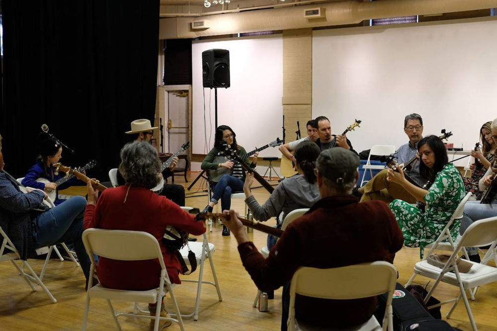 A photograph of a group of people sitting in a circle of folding chairs, each holding a banjo and looking down at the instrument in concentration.