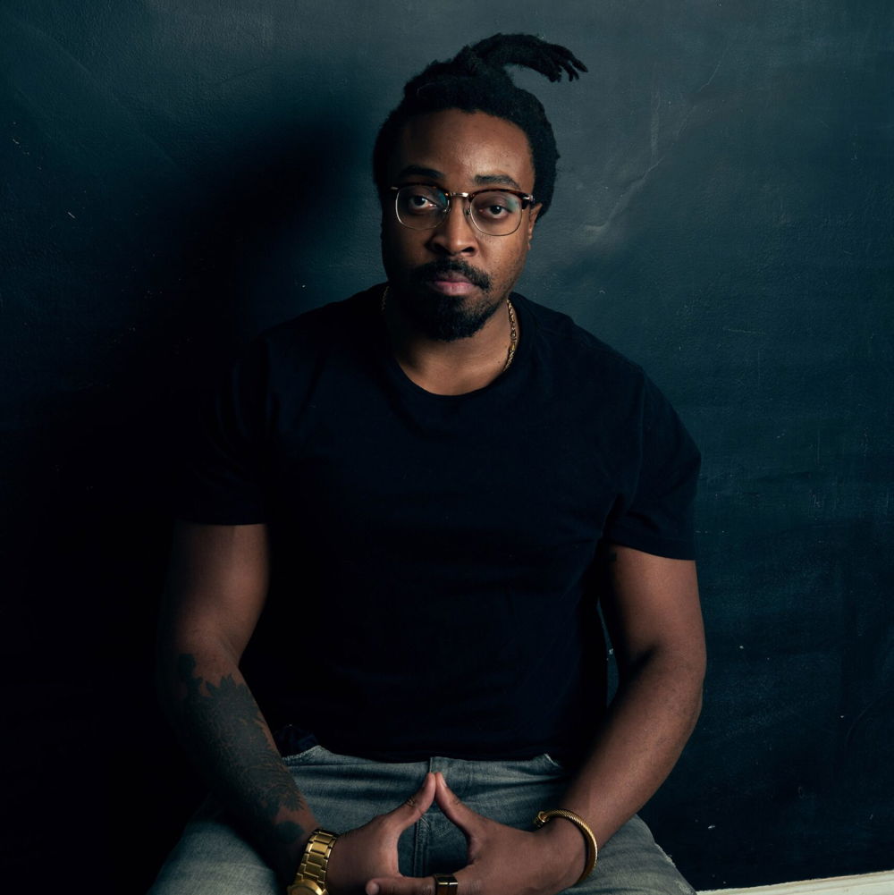 Olalekan is seated against a dark chalkboard background with his hands clasped together in his lap. He is wearing a black t-shirt and dark blue jeans.