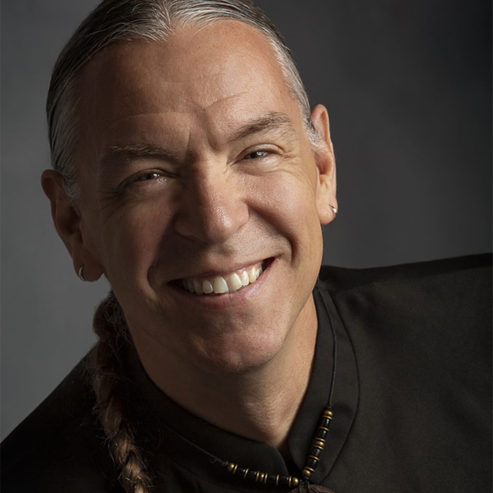 A Chickasaw man with braided hair smiles brightly. He is wearing a dark suit jacket and a traditional shell-carved medallion.