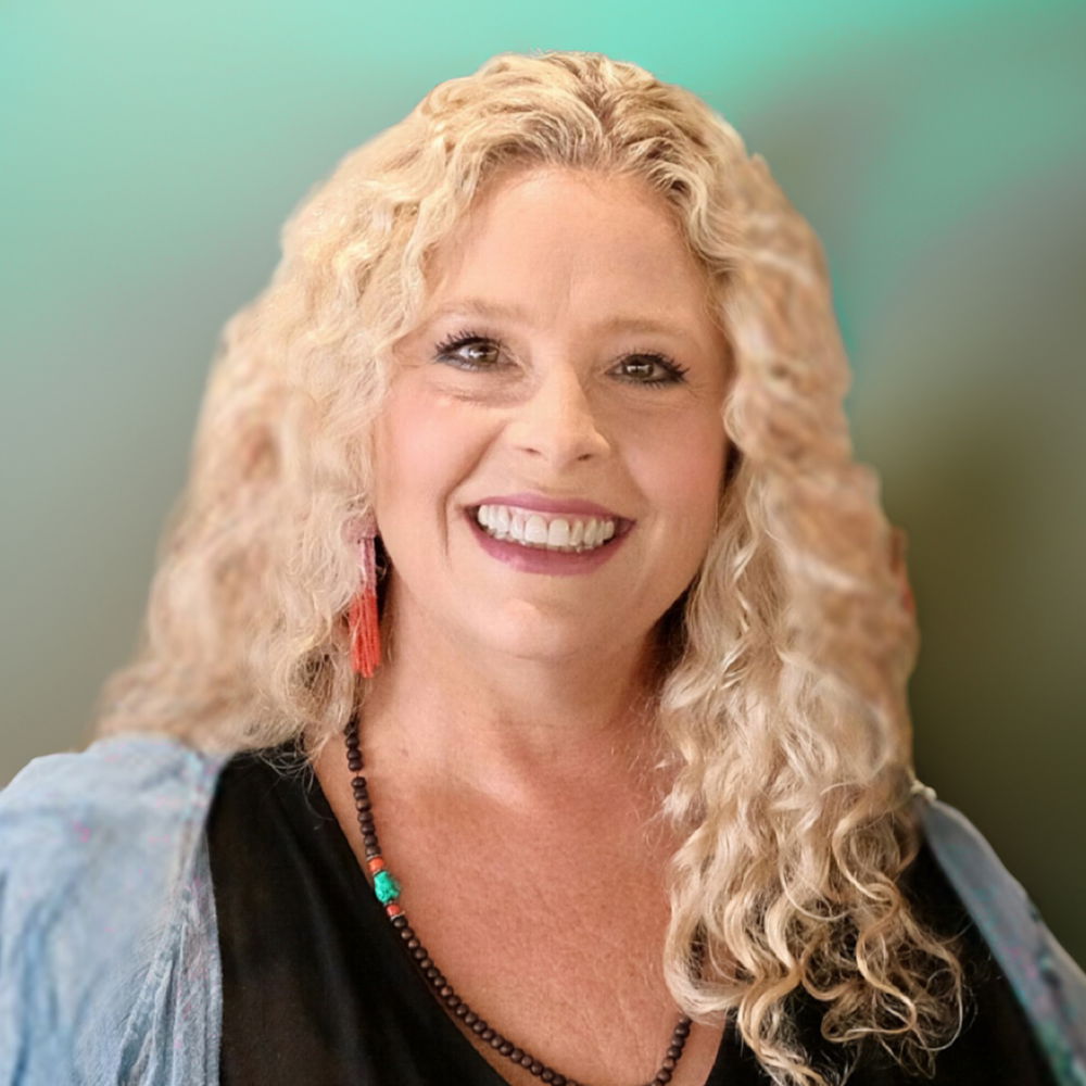 Beth, a woman with long curly blonde hair, faces forward and smiles directly at the camera. She is wearing a dark shirt, beaded necklace, and fringed earrings.