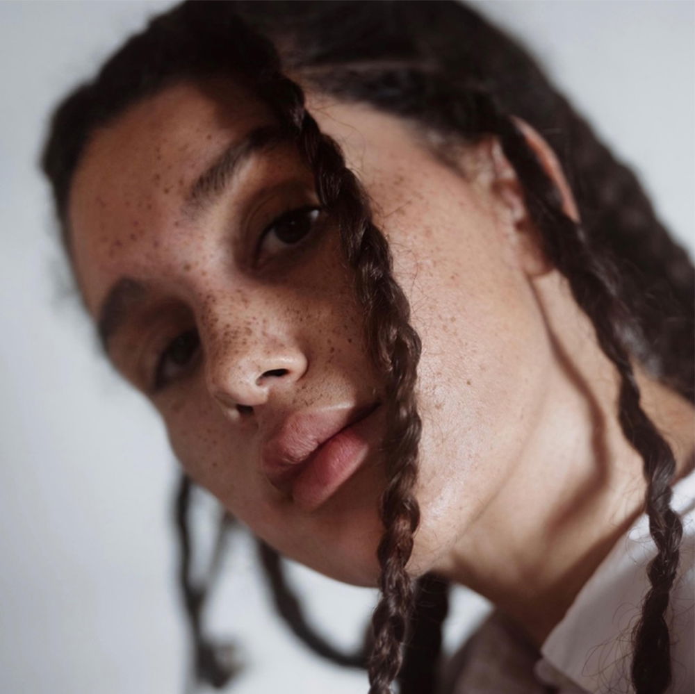 A woman with freckles and braids, looking at the camera with her head cocked to the right of the frame.