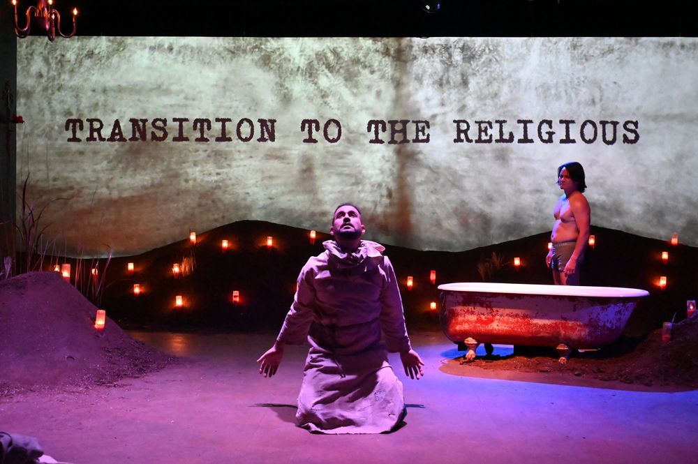 A man in a plain robe kneels on a stage under purple lights, facing the camera with his arms outstretched. Behind him is a large projection that reads, "Transition to the religious." A second figure stands in the background next to a bathtub, surrounded by dozens of small candles on the ground.