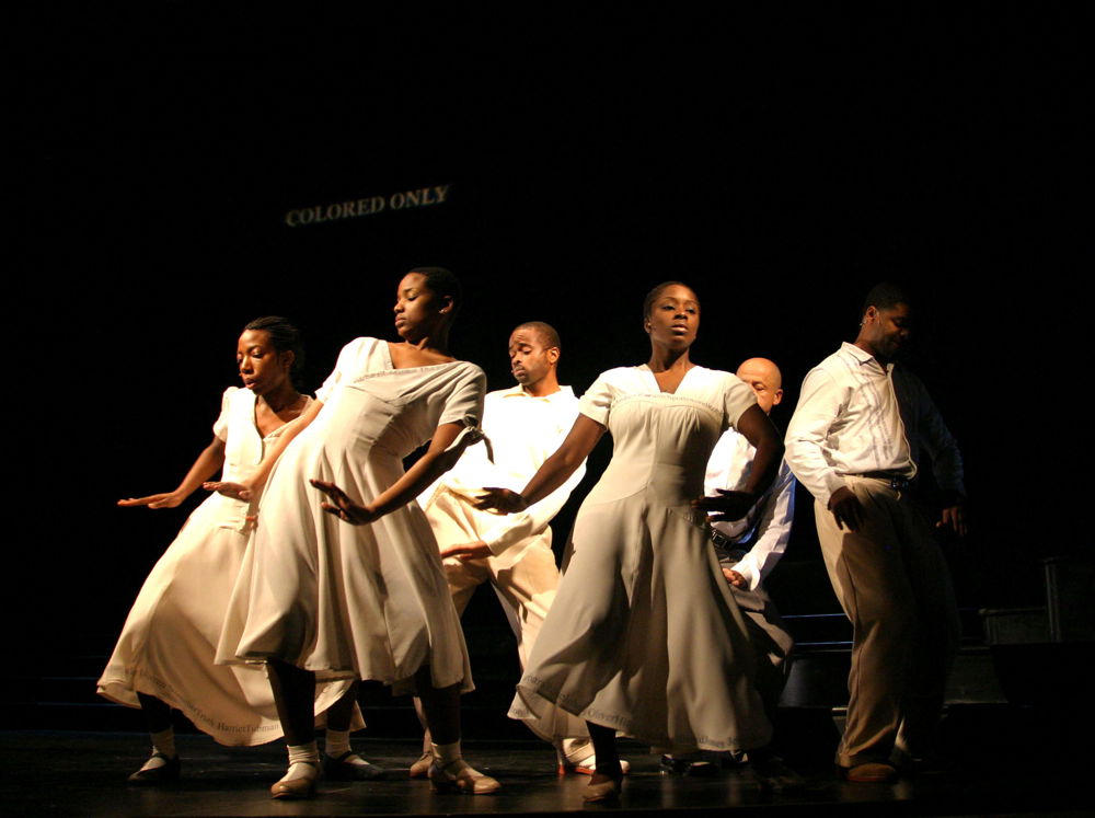 A group of black dancers dressed in white move onstage underneath a sign that reads "colored only."