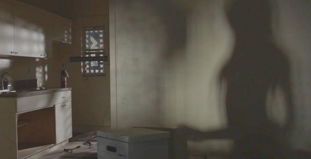 A person’s shadow stretches up the blank wall of an abandoned room. A sink, barred window, and fire hydrant are visible just beyond the wall.