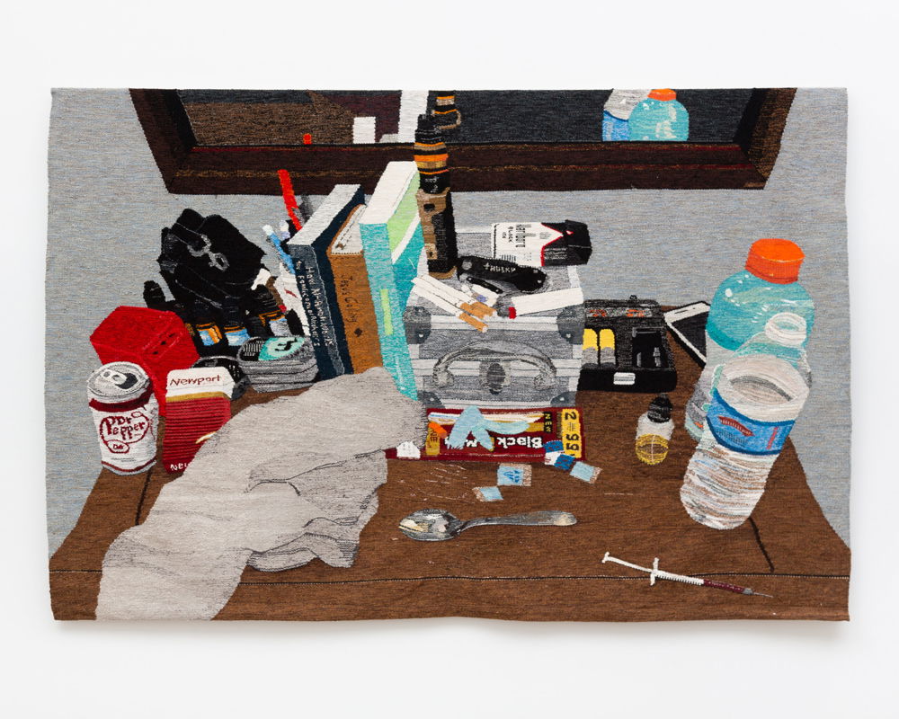 Hanging on a wall is a wool tapestry of a still life of a personal bureau covered in cigarettes, bottles of water, Gatorade, Dr. Pepper, and drug paraphernalia. It has a stack of books containing Jesus Calling and How Al-Anon Works as well as cigarillo packs, many vape liquid containers, and an iPhone.