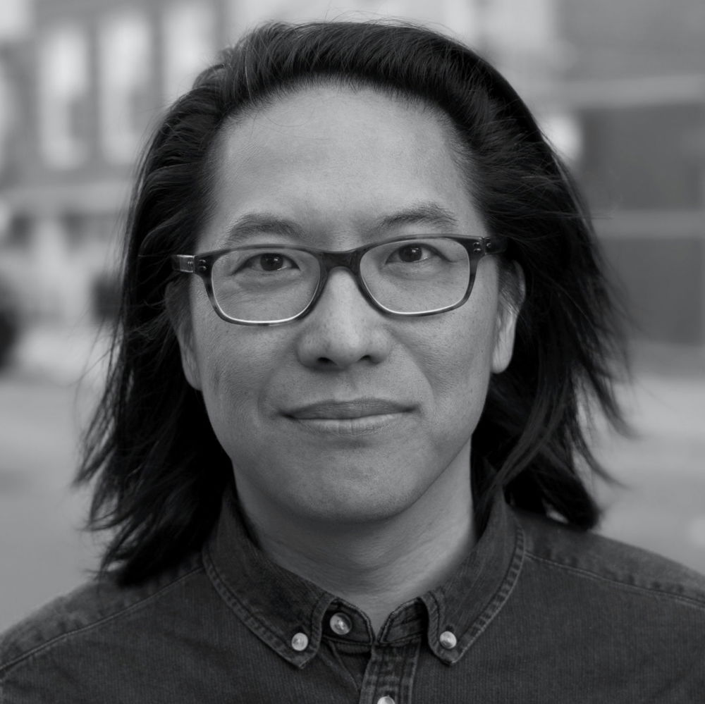 Stephen, an Asian-American man with shoulder-length black hair, glasses, and a dark shirt, looks into the camera.
