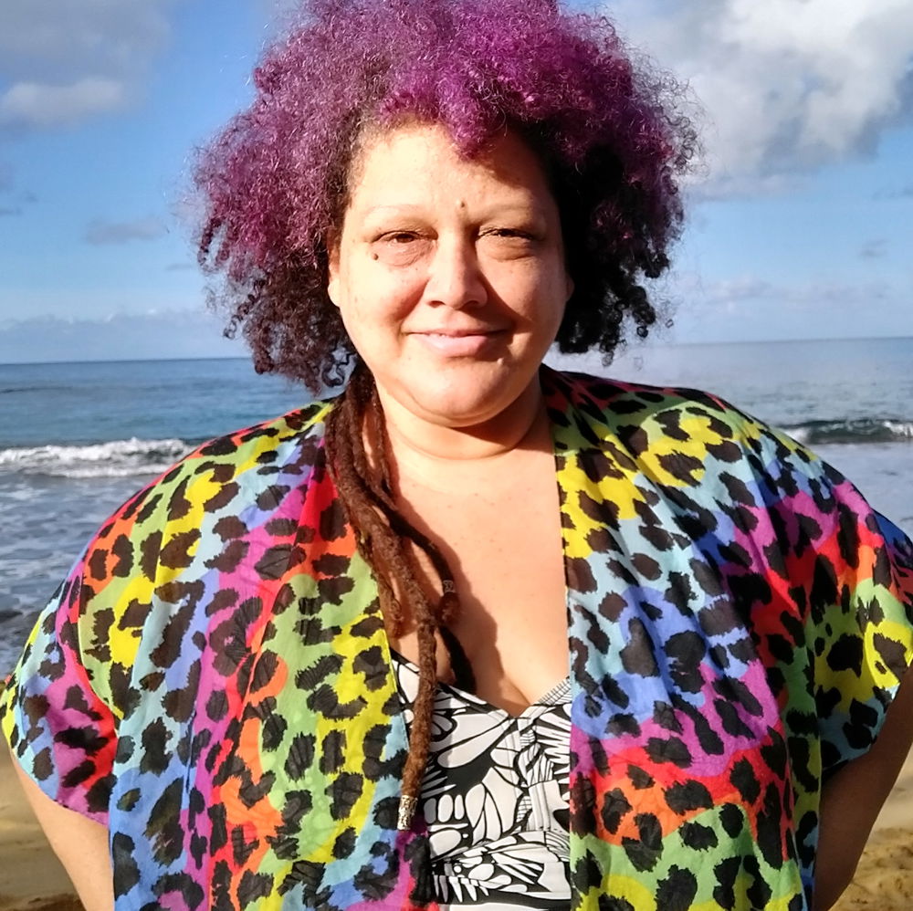 Macha Colón, a light-skinned Afro Puerto Rican with purple dyed curly hair, smiles at the camera. She stands at the beach with the ocean behind her.