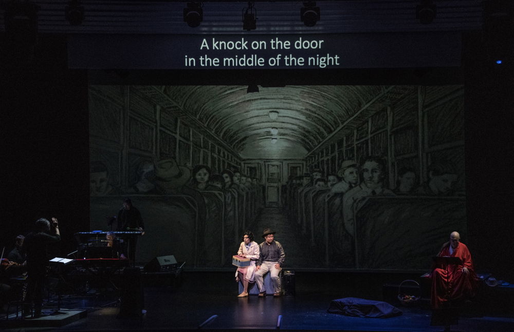 Two people crouch center stage, as though in flight. A monitor at the top of the stage reads “A knock on the door in the middle of the night.” A stylized black-and-white illustration of a train car filled with solemn passengers forms the stage’s backdrop.