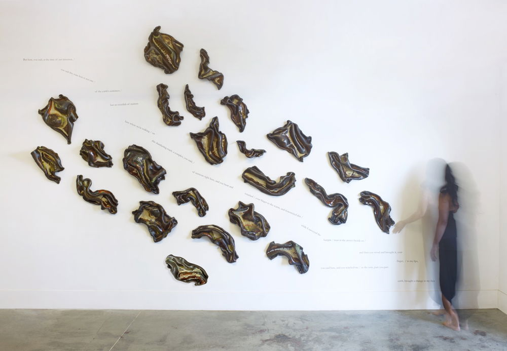 A grouping of abstract ceramic sculptures scattered across a white wall. Each individual sculpture resembles torn scraps of brown fabric or paper that have been burnt around the edges until they wave and curl. Lines of text divide the group of sculptures in half. A blurry figure appears standing to the right of the sculptures.