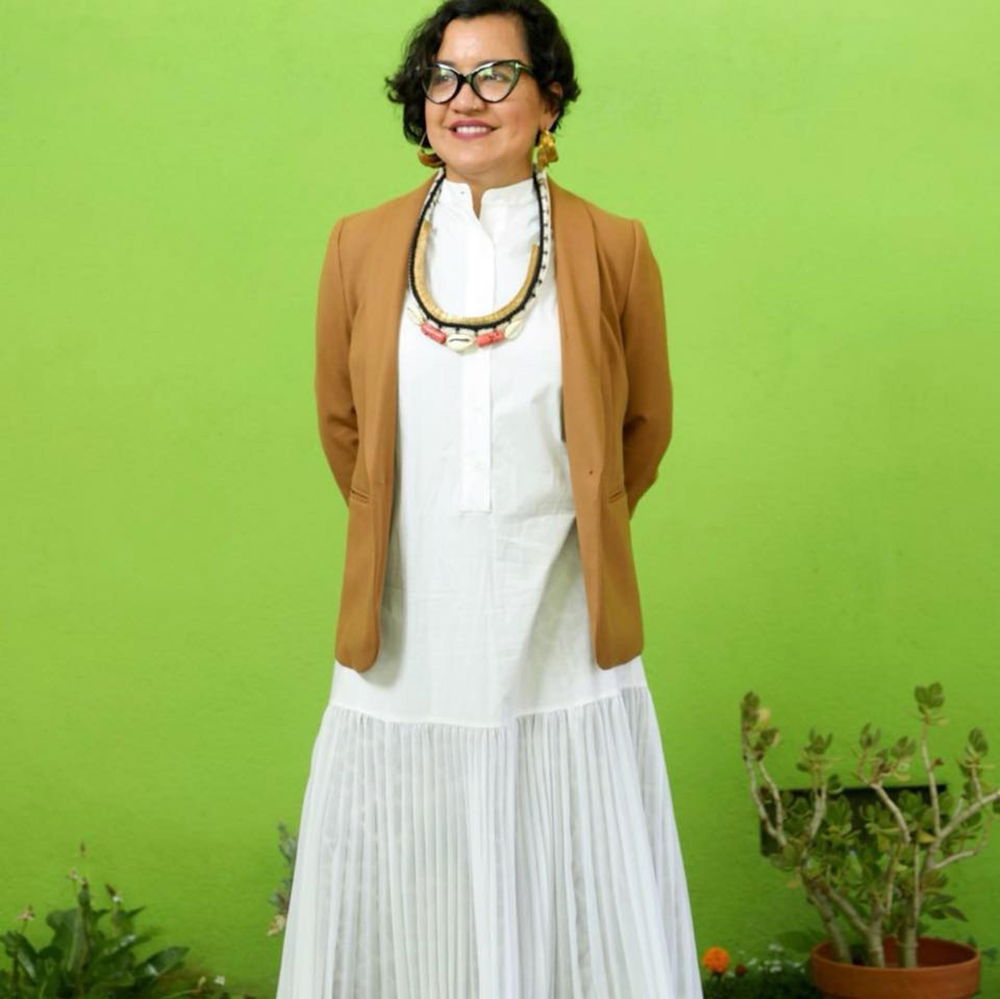 Martha poses in front of a lime green wall in a long white dress, caramel blazer, cat eyeglasses, and jewelry. She is a Chicana woman with tan skin, short black hair, and a warm smile.