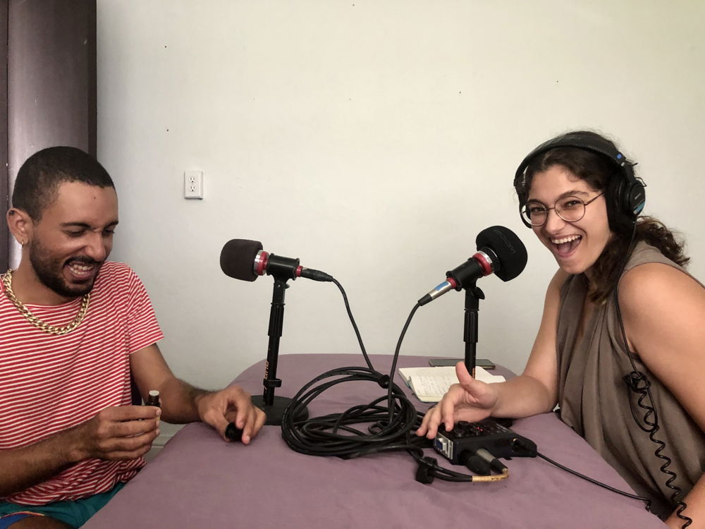 Two people laugh and smile on either side of a lavender table in front of microphones. The person on the left has medium-brown skin, close-trimmed hair and beard, and wears a red-and-white striped shirt. The person on the right has long dark hair pulled back into a low ponytail and wears thick black headphones, wire-rimmed glasses, and a gray sleeveless shirt.
