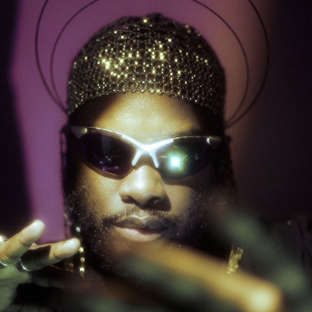A close-up of a person's face wearing black shades and a golden headdress with a golden halo. They have medium-brown skin and a neatly trimmed beard. Their face is partly obscured by their outstretched hands.