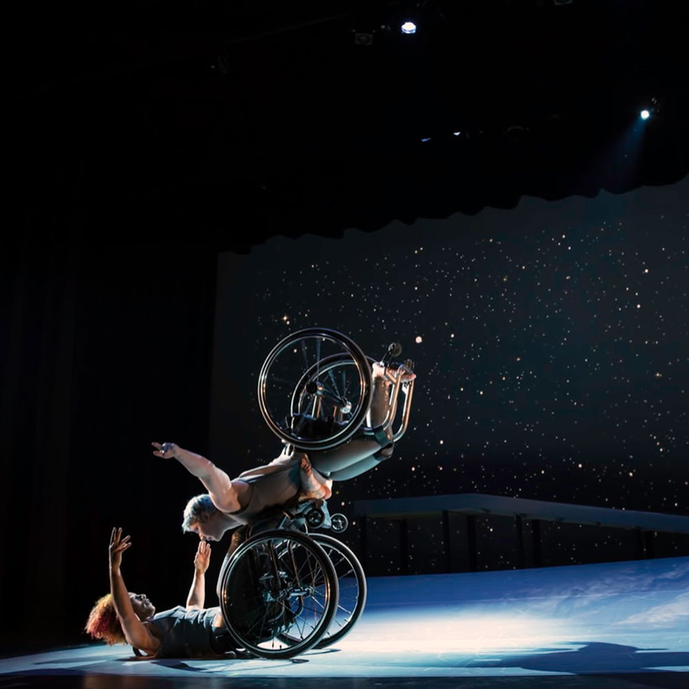 Venus balances on the footplate of Andromeda’s wheelchair, arms spread wide and wheels spinning. Andromeda opens her arms wide to receive her in an embrace. They make eye contact and smile. A starry sky fills the background, and moonlight glints off their rims.