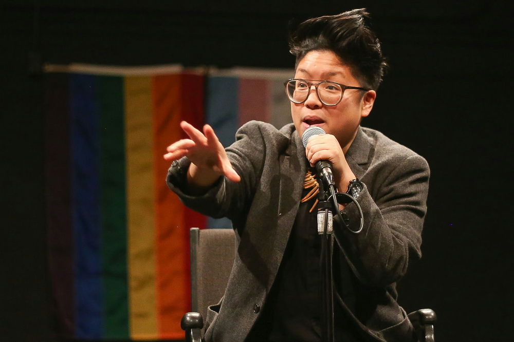 Kay, a brown, round queer, performs at a microphone with transgender and rainbow flags in the background. They wear a gray blazer and glasses.