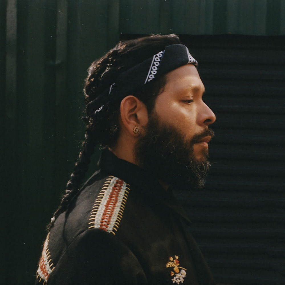 A bearded person stands in profile looking off camera. His long, dark hair is woven into two braids. He wears a black bandana around his forehead and a black shirt with colorful embroidered accents.