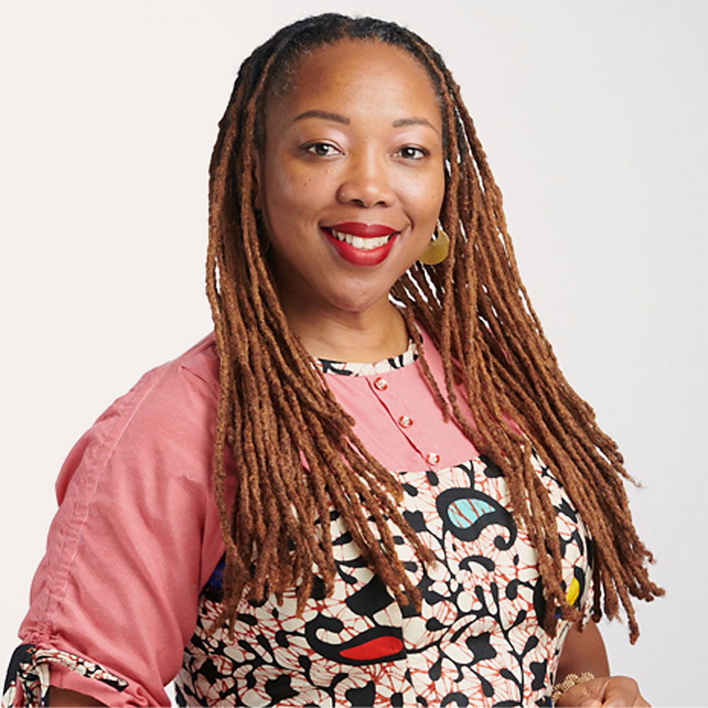 A woman of African descent smiles. She has copper-colored locs past her shoulders and is wearing red lipstick and a blouse with a pink-and-white Ankara print.
