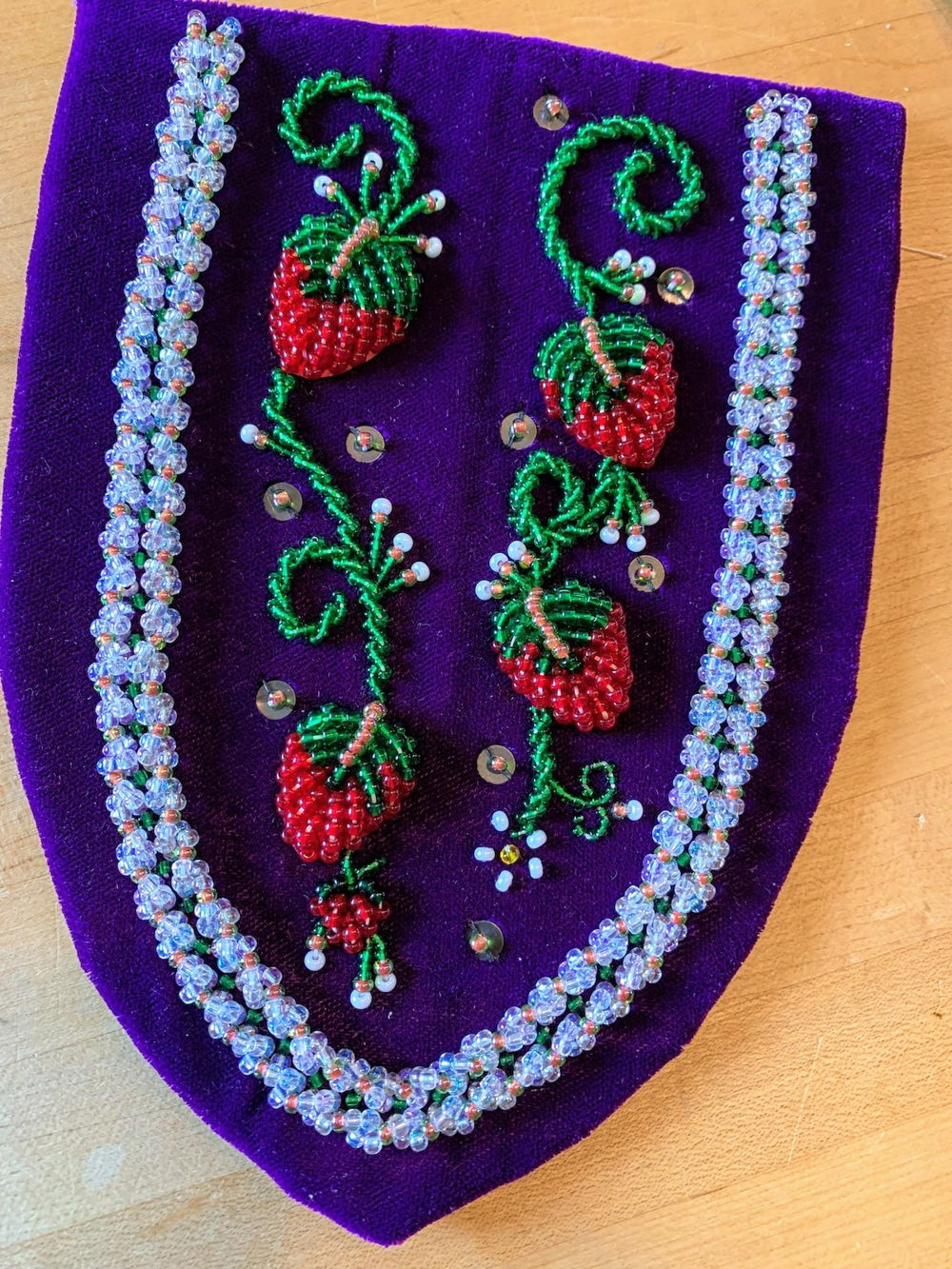 [ID: A semicircle piece of purple fabric embroidered with intricate beadwork of red strawberries, white flowers, and green leaves.]