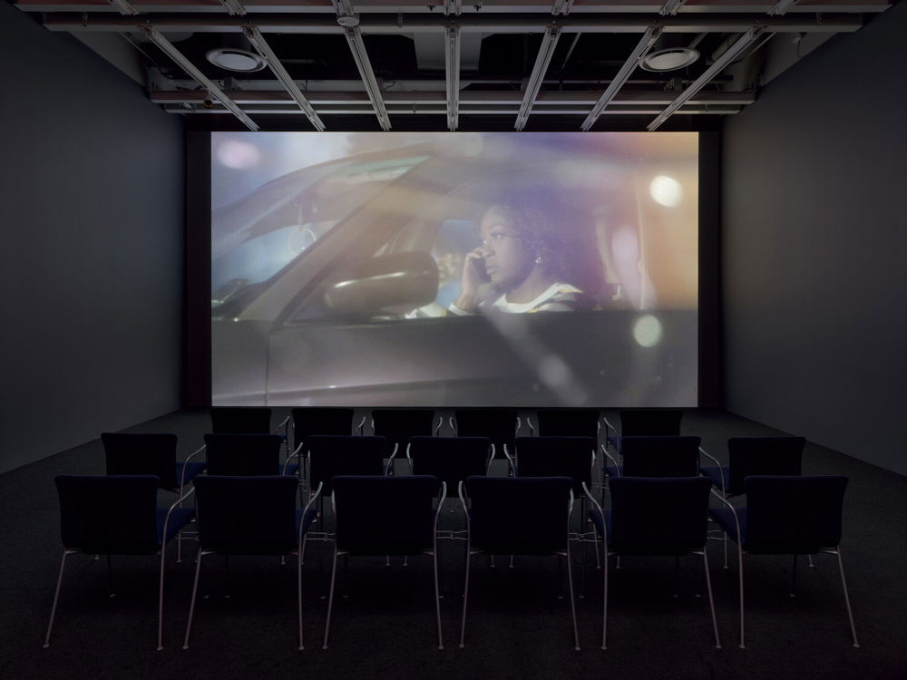 A small dark theater with rows of empty chairs oriented towards an image projected on a back wall. The image is of a Black woman talking on a cell phone in the driver's seat of her car. The image is seen from outside of the vehicle. The lighting is dreamy and soft.