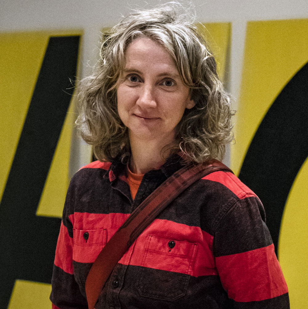 Sharon, a middle-aged white queer woman with wavy brown and gray hair, stands in a gallery space in which an artwork is being installed. She is wearing a long shirt with wide black and red horizontal stripes and a camera strap diagonally across her chest. She looks at the camera and is smiling; her right hand is in her pocket, and her left forearm is behind her back.