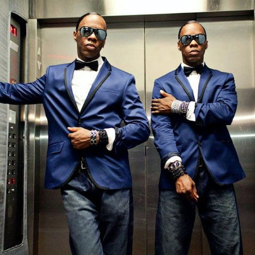 A portrait of two seemingly identical men in an elevator interior. Both men are Black with short hair and matching attire. They both wear large reflective glasses, blue tux jackets trimmed in black, black bow ties, white dress shirts, and blue jeans. The man on the left rests his extended right arm on the elevator wall and his left hand at his navel. The man on the right, holds his right bicep. Both men regard the viewer with cool sartorial looks.