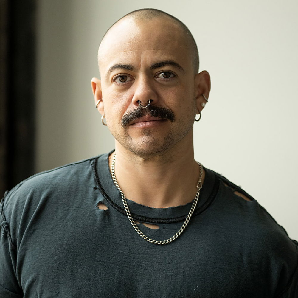 Headshot of an artist in his studio. He looks directly at the camera and wears a gray blue T-shirt that is torn along some of the seams. He has light tan skin, a shaved head, and a thick brown mustache.