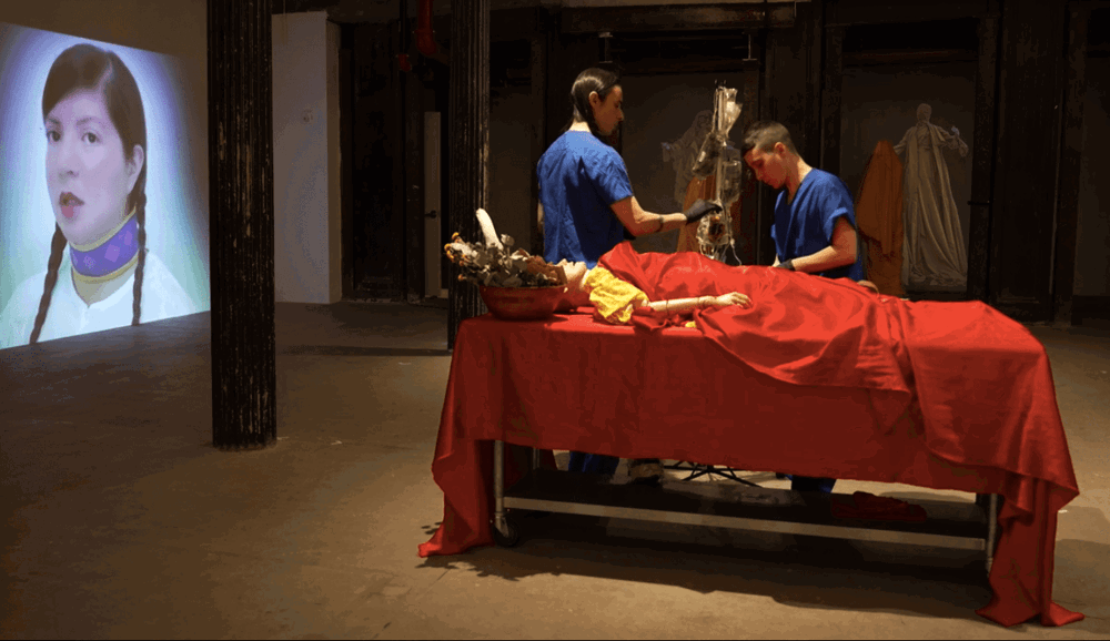Two performers in blue medical scrubs hang an IV bag. In front of them, a medical mannequin lays on a red table covered in a red sheet. In the background of the set is a projection of a woman's face and two statue-like figures draped in yellow.
