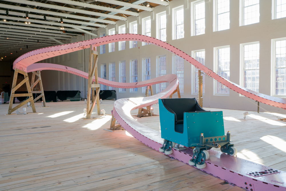 A sculpture placed inside a large gallery space that resembles a roller coaster. The thin, pink track curves and ripples around the spacious modern interior, upheld by wooden frames. In the foreground sits the roller coaster car, a modest two-seater painted teal.