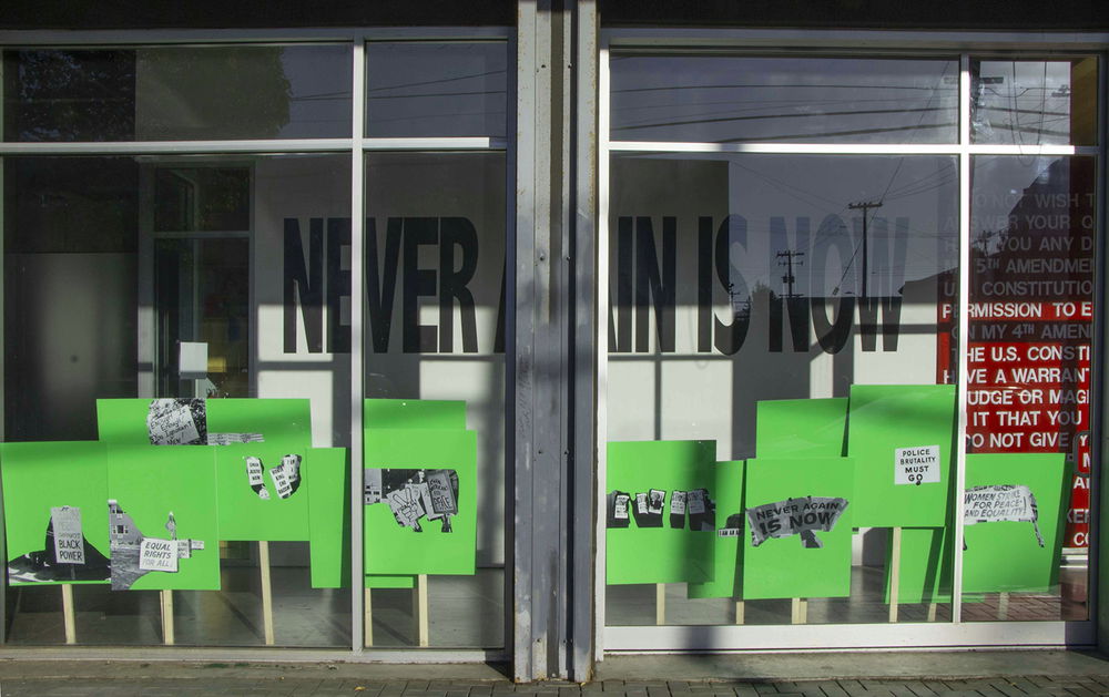 Fourteen neon green posters on wooden pickets of varying lengths lean against the glass inside of a storefront window. On each poster is a printed black-and-white image of protest posters from different eras, including ones that say "Police Brutality Must Go," "Yellow Peril Supports Black Power," "Enough Is Enough! You Ignorant Men!," and "Equal Rights For All." On the gallery wall behind the pickets, large black vinyl text reads, "NEVER AGAIN IS NOW."