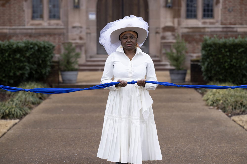 A dark-skinned woman dressed in a white dress and a white hat that features white mesh detailing. She holds a knotted blue fabric rope and poses in front of a large, elegant building.