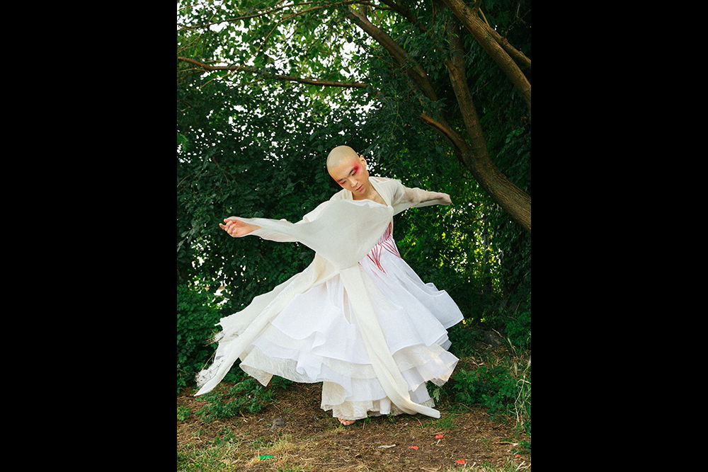 [ID: Eddy is wearing a long robe and layered dress made of sheer white fabrics with red accents on the torso. They are dancing in front of trees and green leaves and spinning with their arms extended and eyes looking toward the ground. Red makeup accents each temple.]
