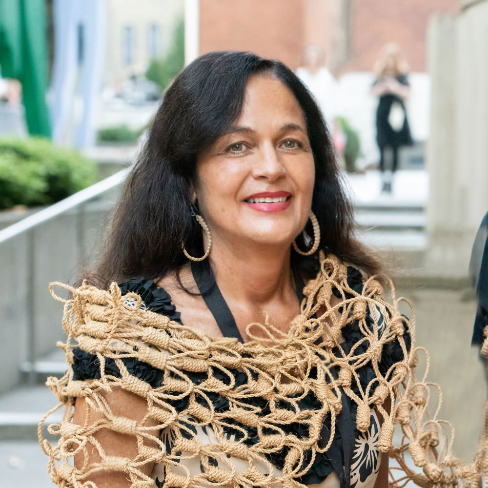 Lulani Arquette, a Native Hawaiian woman wearing an a`ahu `aha, coconut cord wearable piece made by artist Marques Marzan and matching hoop earrings over a black dress, smiles warmly at the camera.