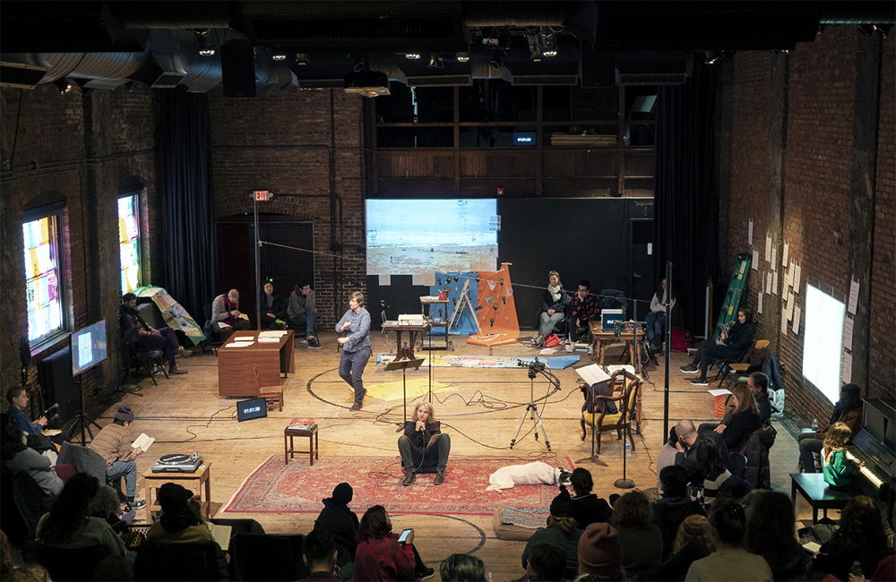 A bright, brick-walled industrial space with an elaborate array of performers, stage hands, and equipment scattered around. An audience sits along the bottom of the image watching.