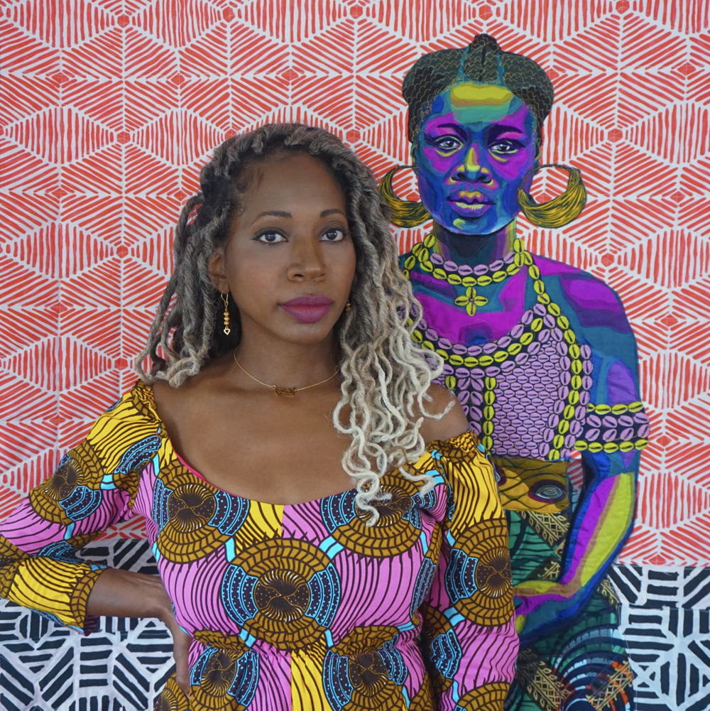 Bisa, an African American woman with long cascading locs, dark lipstick, and a bright printed dress stands in front of her quilt with an equally colorful portrait of an African woman with short, parted hair, hoop earrings, and a halter made of cowrie shells.