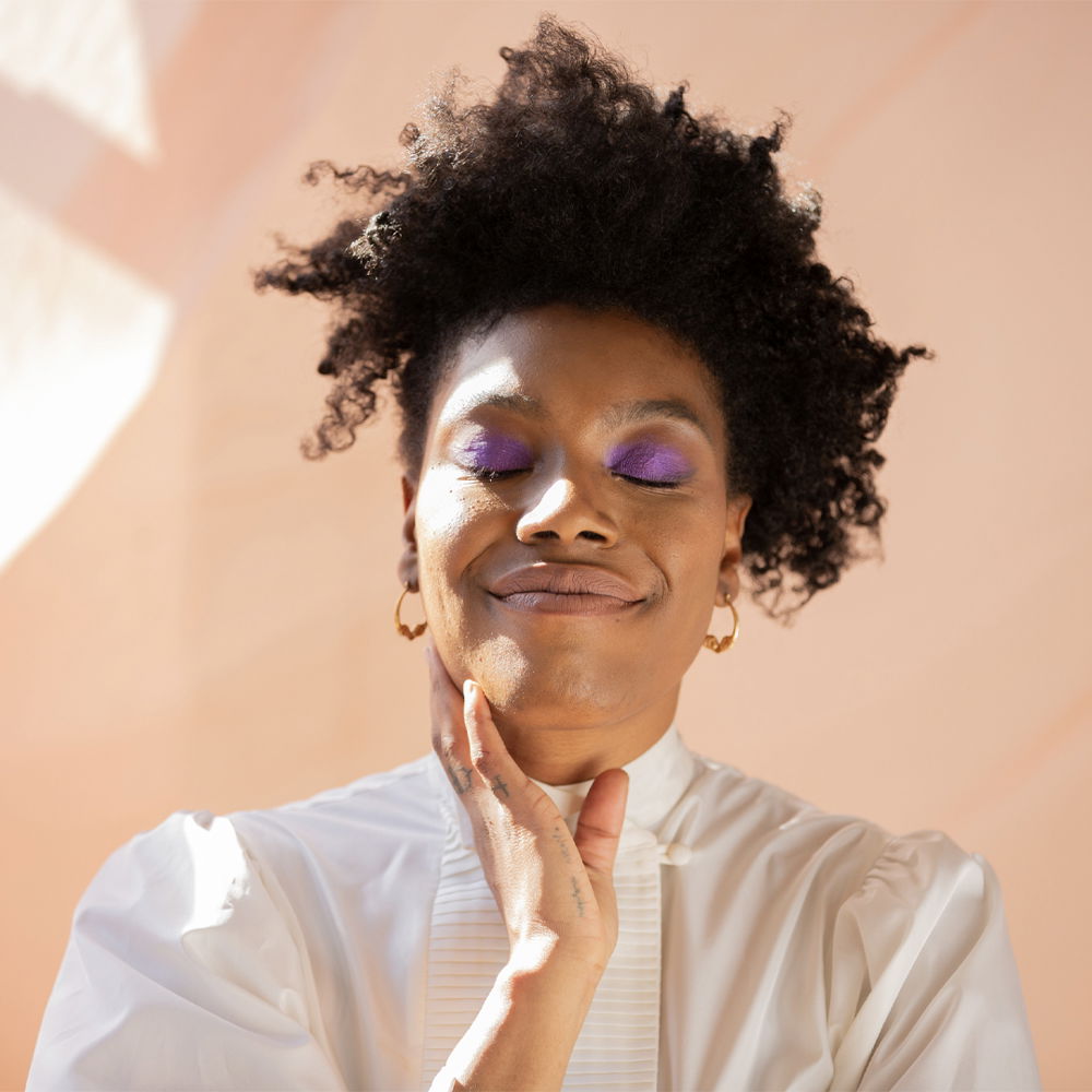 A portrait of a Black woman with an asymmetrical afro, standing in front of an apricot-colored wall. Her eyes are closed and adorned with purple eyeshadow. She appears to be breathing in, mid-inhale. Her fingertips lightly touch her neck, her face glowing, and her lips spreading into a big smile, giving a look of both serenity and joy.