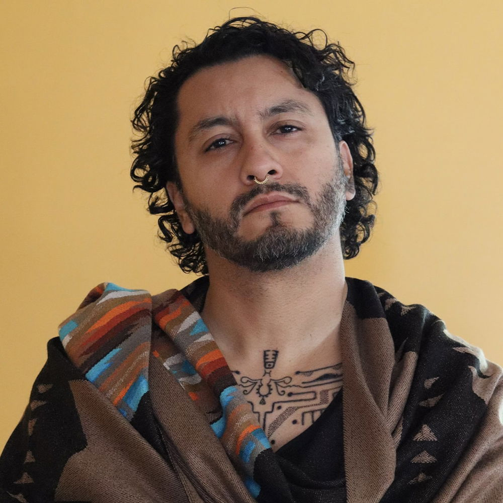 A headshot of a Latinx person of Mexican-American descent wearing a serape poncho posing in front of a yellow wall. His face is slightly tilted back, and he is wearing a gold septum piercing and staring directly at the viewer.