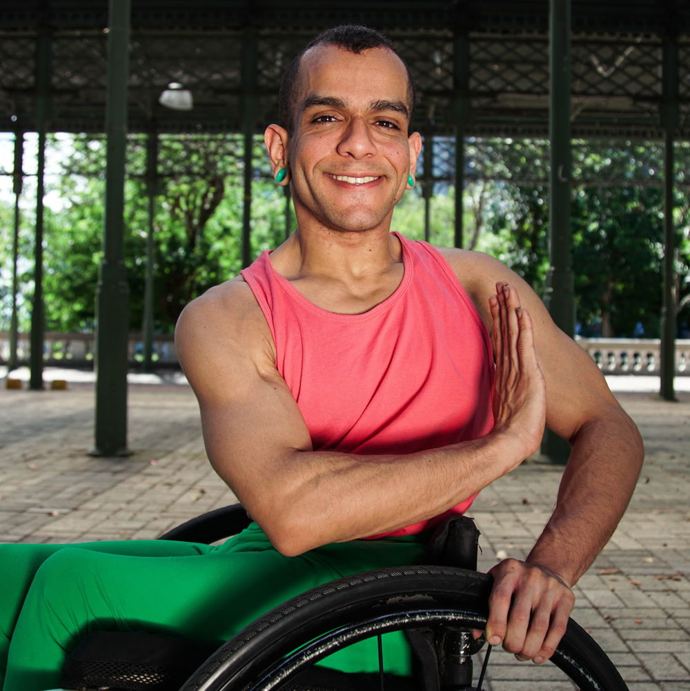 JanpiStar, a wheelchair user with light brown skin, close-cut dark hair, and an athletic build, smiles and poses with their right upper limb in front of their chest, hand flexed. They wear neon green gauge earrings, a coral tank top, and green leggings.