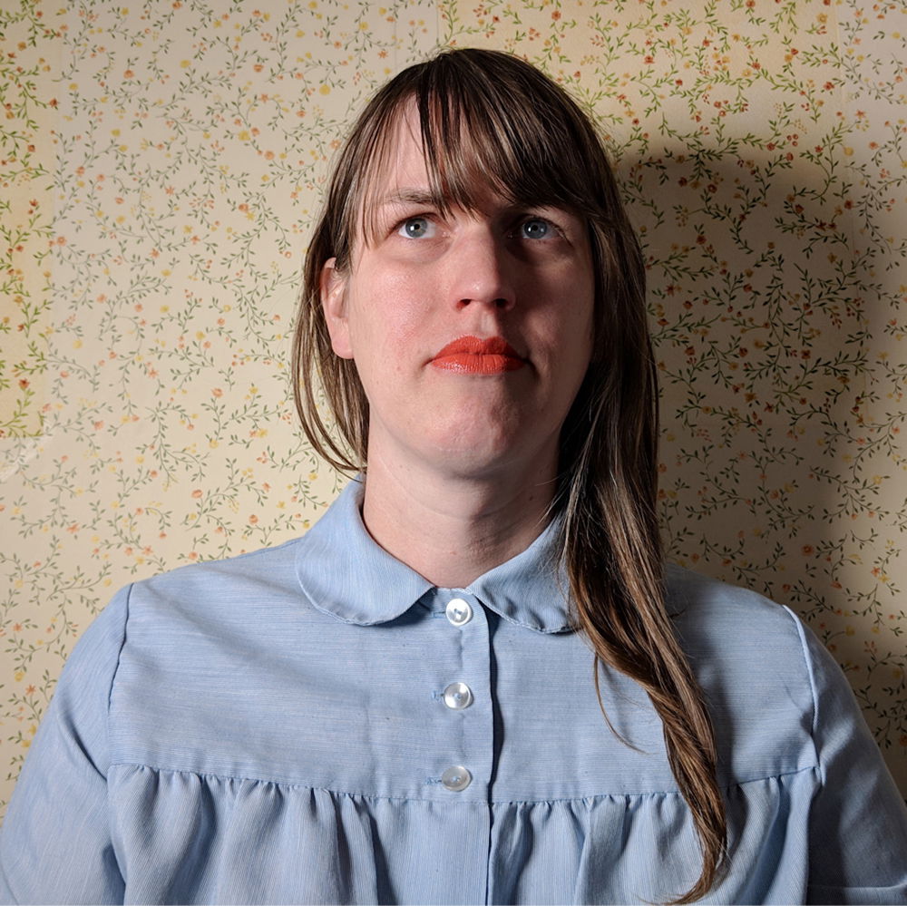 A woman photographed from the shoulders up wearing a blue collared shirt and red lipstick with long hair combed over the left shoulder. She is positioned in front of mostly beige floral wallpaper drawing by visual artist Mara Baldwin.